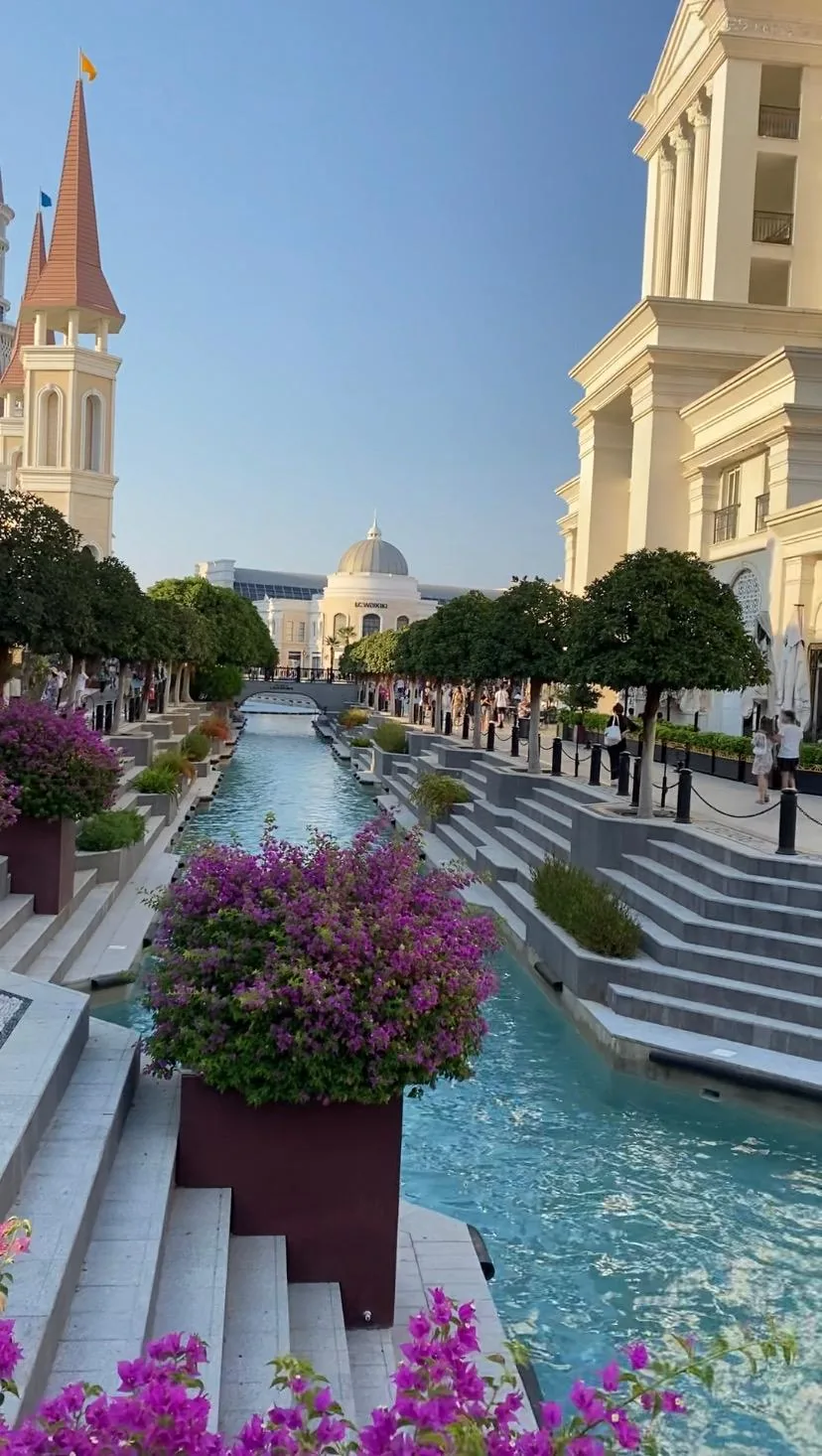 A turquoise canal with stone steps and pink flowering planters running between white buildings toward a bridge