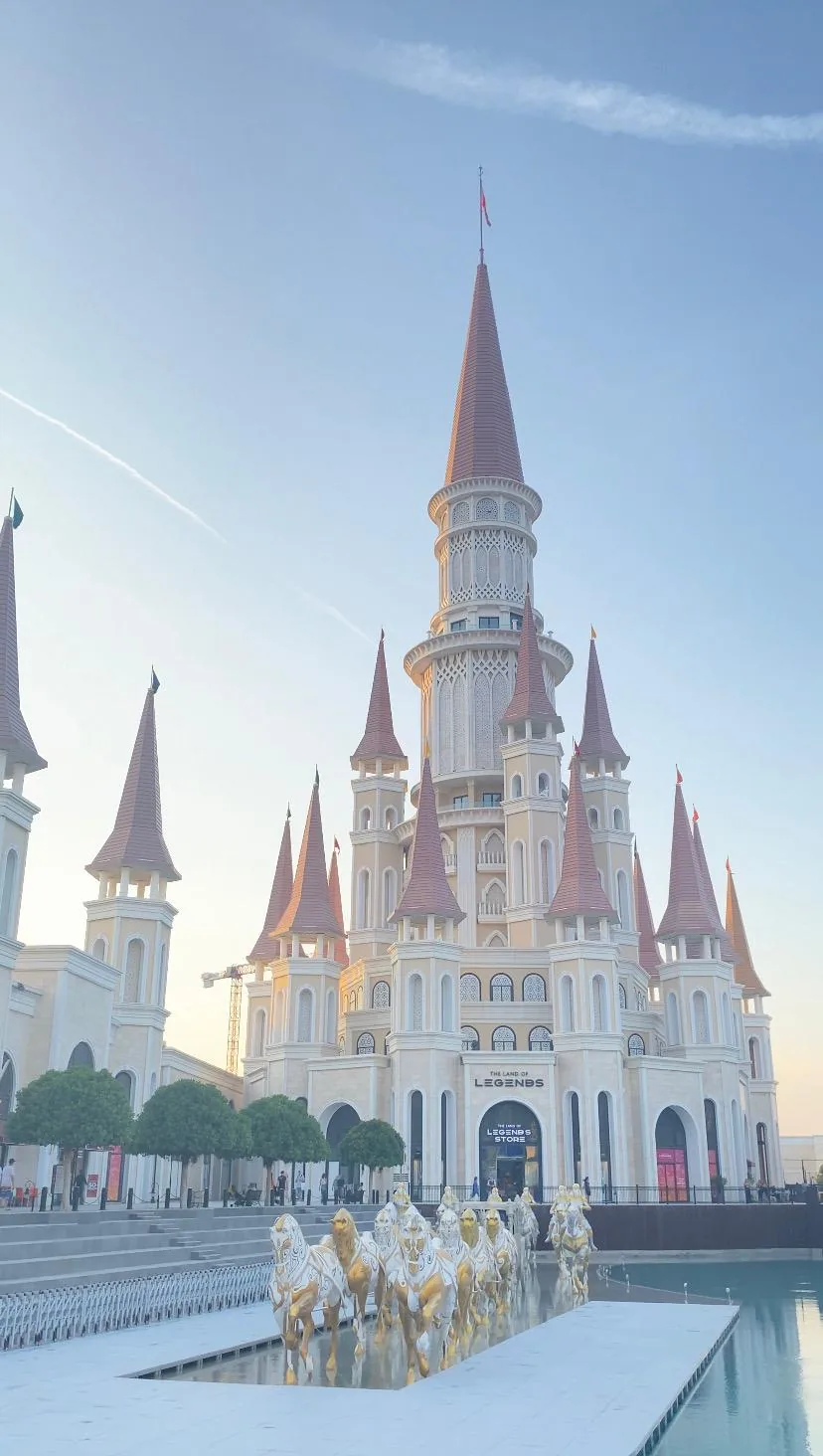 A pale castle with a tall central tower and rust-red spires beyond a pool where gilded horses stand, in soft morning light